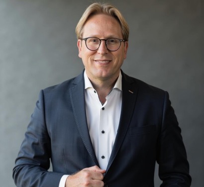 Harold Koenjer in donkerblauw pak met bril, glimlachend voor grijze achtergrond
